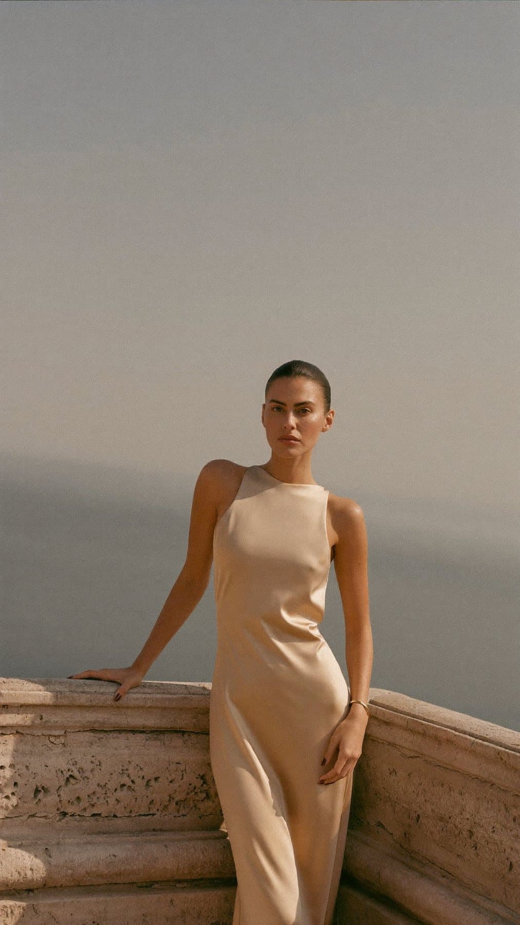 A woman in a flowing sand silk dress on a sunlit limestone terrace above the sea